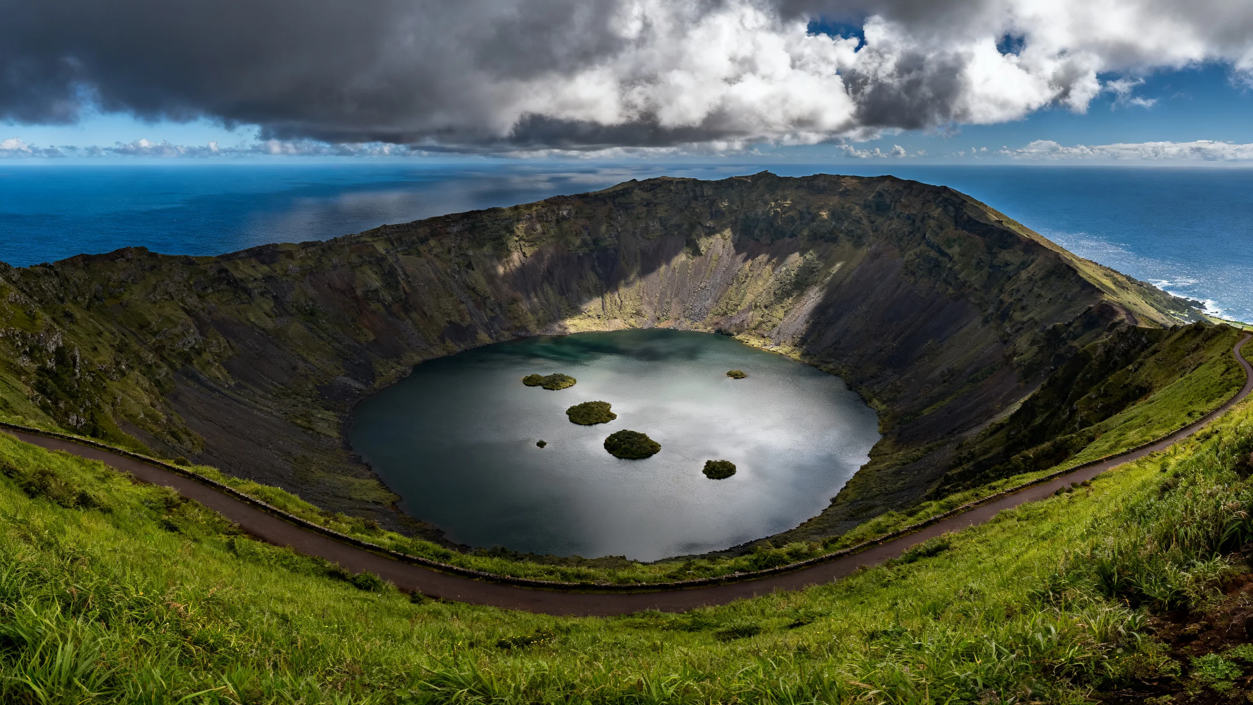 A panoramic view from the rim of the Caldeirão of Corvo, Azores, looking down into the 2 kilometre wide volcanic crater, with the small Lagoa do Caldeirão lake at the centre and several small islets visible in the lake, the rim path winding around the green edge, dramatic broken clouds overhead