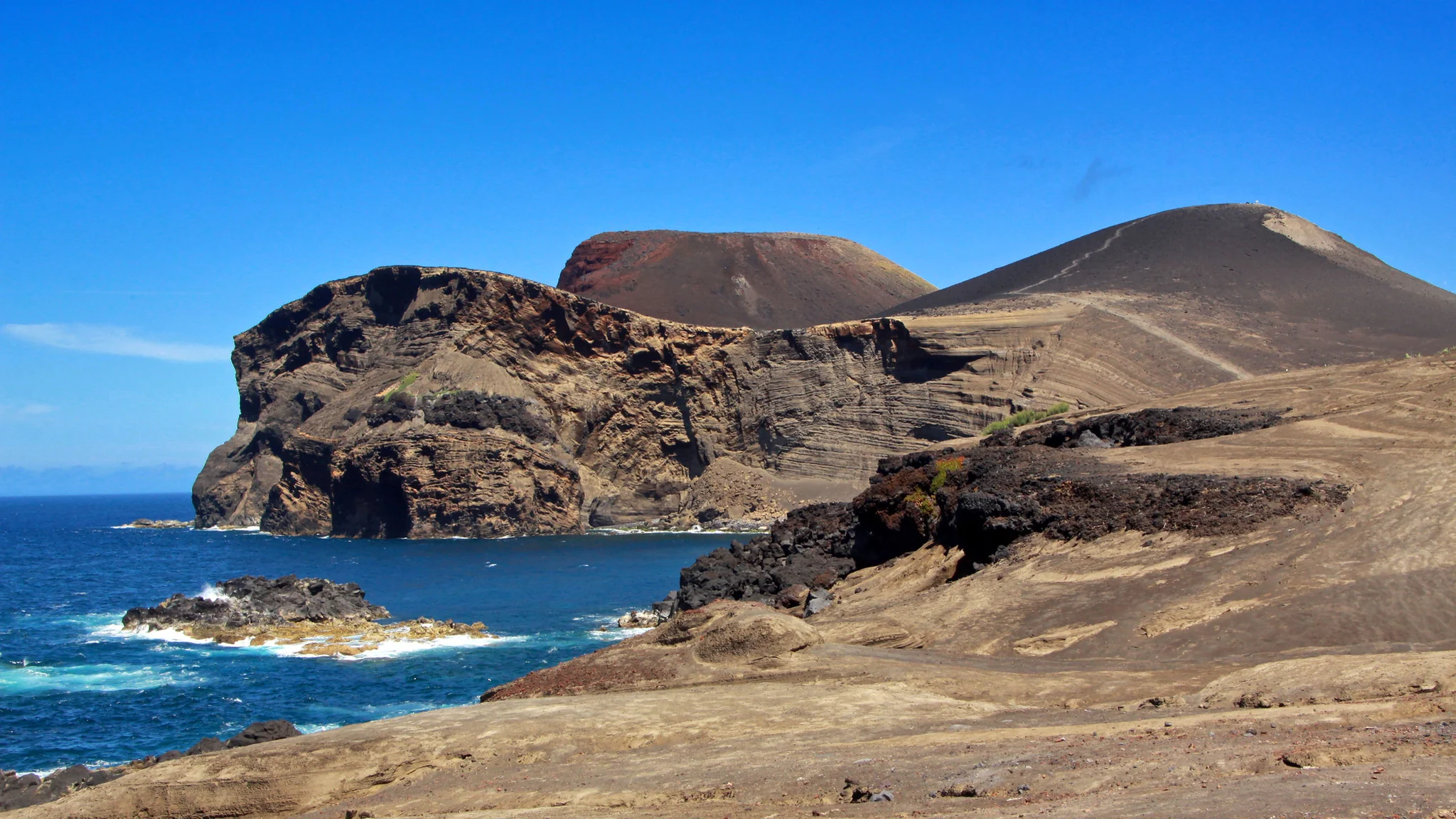 The Capelinhos volcanic peninsula on the western tip of Faial, Azores: red-brown cliffs of volcanic scoria meeting the Atlantic, the dark cone of the 1957 to 1958 eruption rising in the background under a clear blue sky